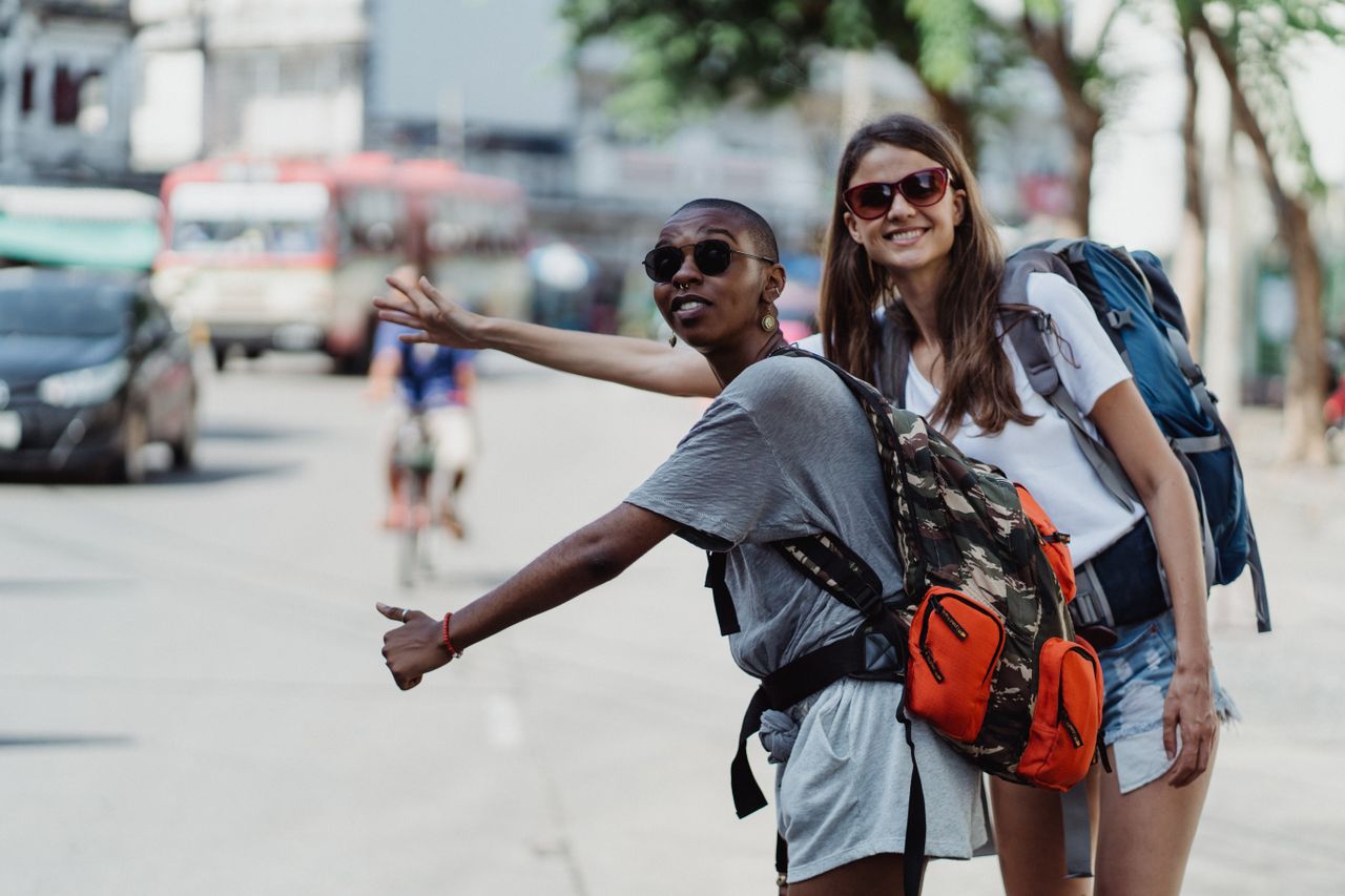 Two women with backpacks hitchhiking in a lively city street, embracing adventure.